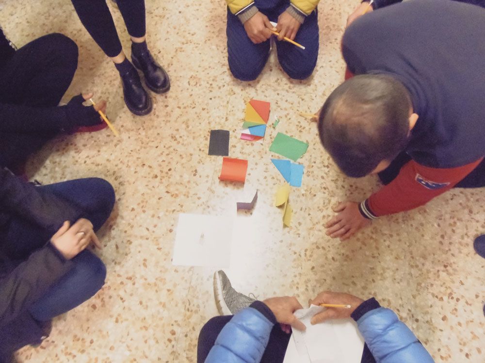 L'immagine mostra sei persone disposte in circolo intorno a dei campioni di colore; le persone sono viste dall’alto, tre sono sedute e si vedono solo le ginocchia e le mani; una è in piedi e si vedono solo i piedi, l’ultima persona è inginocchiata e si vede la nuca e la schiena. Due persone hanno una matita in mano.