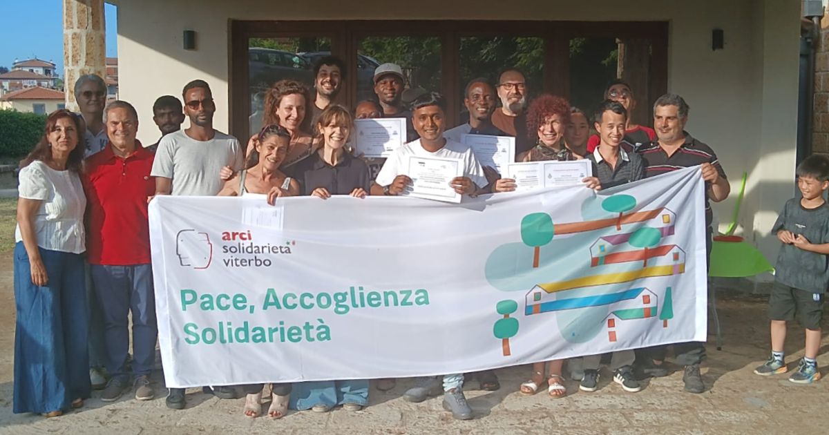I volontari, le operatrici e gli operatori reggono uno striscione di Arci Solidarietà Viterbo con la scritta Pace, Accoglienza, Solidarietà e un disegno colorato con case, alberi e strade