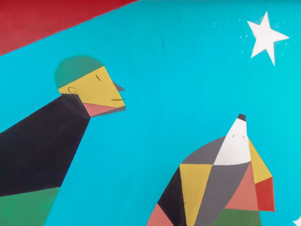 Un particolare di uno dei murales: su un fondo azzurro chiaro due figure nel lato basso osservano una stella nell’angolo in alto a destra dell’immagine. Le figure sono dipinte in modo semplificato con campiture di colori pieni di forma geometrica. La figura umana di sinistra parte dall’angolo in basso a sinistra e è inclinata verso destra; la seconda che parte dal lato basso dell’immagine rappresenta un animale.
